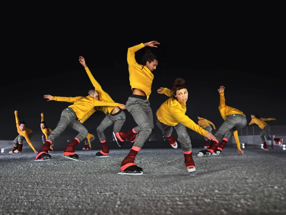 A group of ice dancers, all dressed in yellow shirts and grey pants, in various positions on the ice.