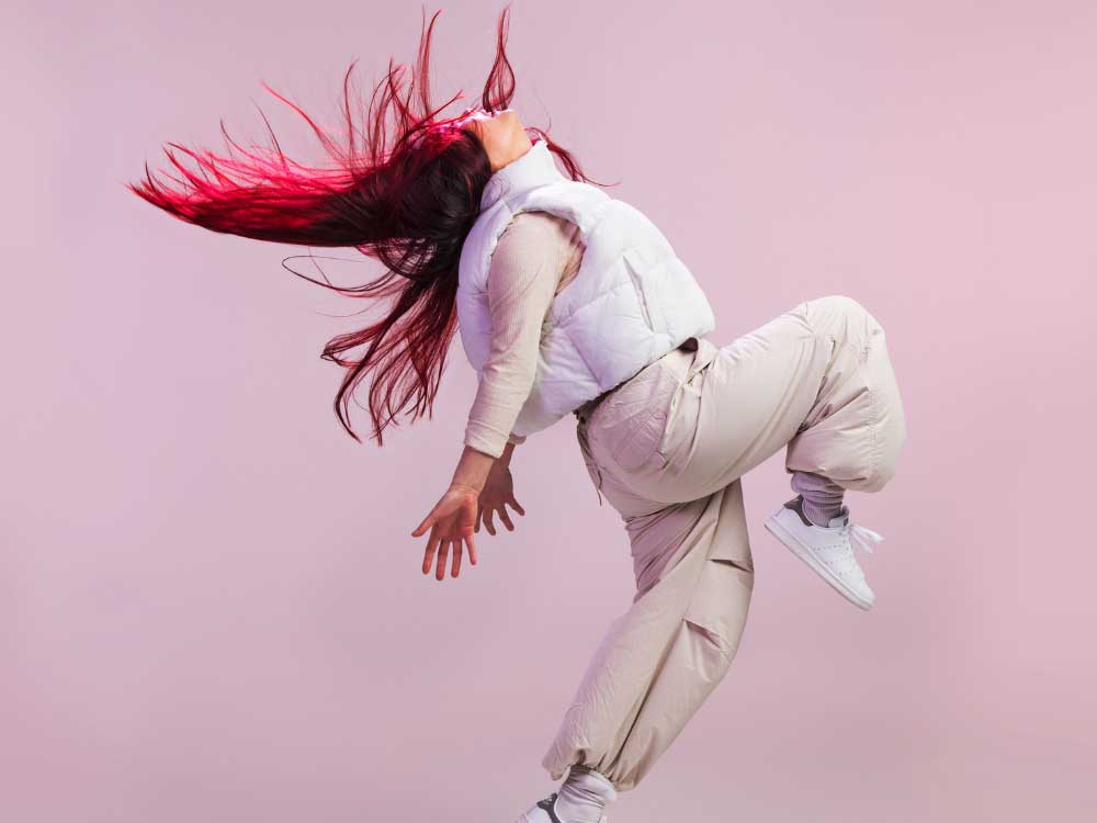 A dancer with long pink hair wearing beige cargo pants, a beige long-sleeve and a white puffer vest is in a dance pose, kicking one leg up in a pike position and pushes their arms behind their torso as their head and chest tilt back. 