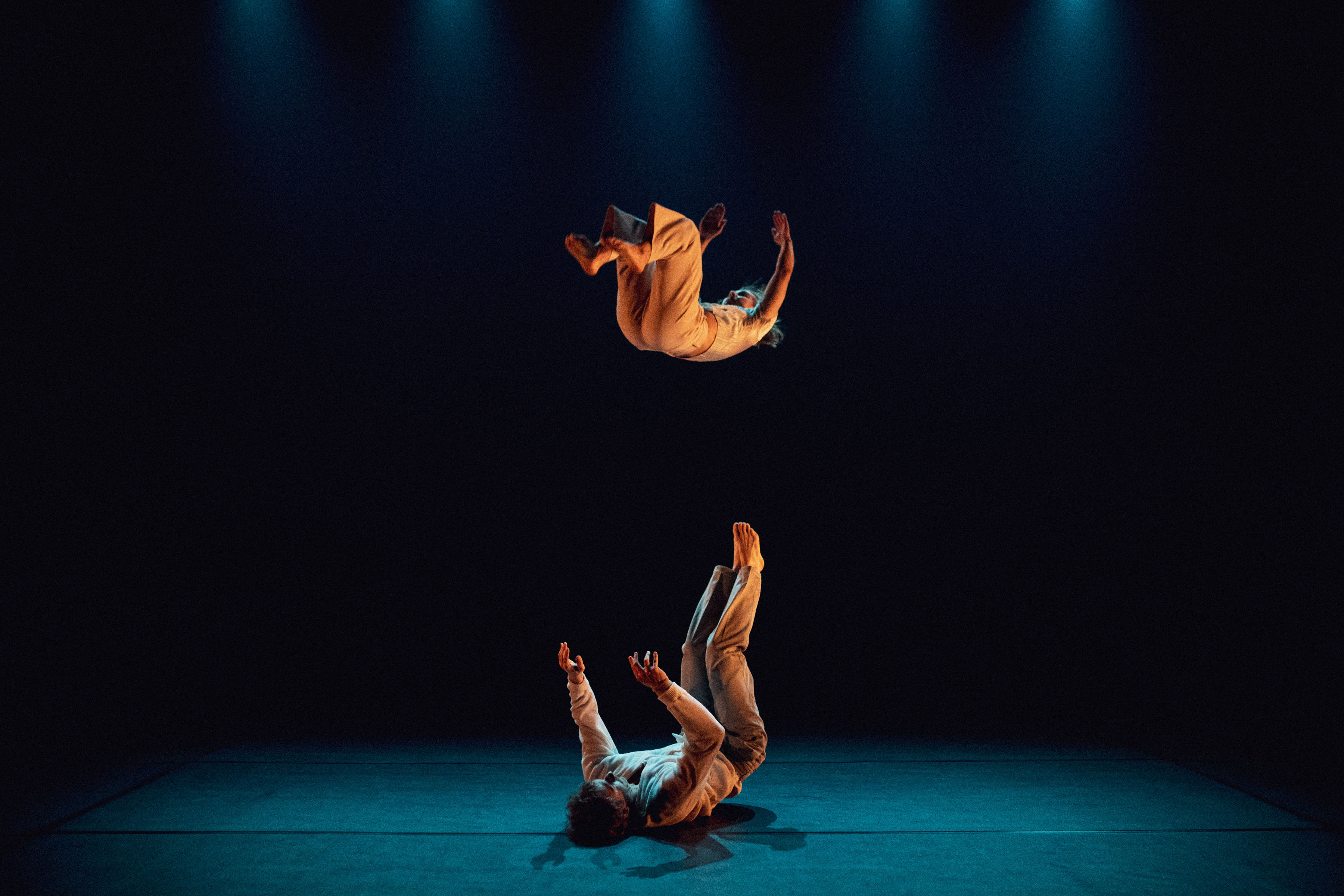 On a stage, a dancer is on their back on the floor and appears prepared to catch another dancer who is mid-air. 