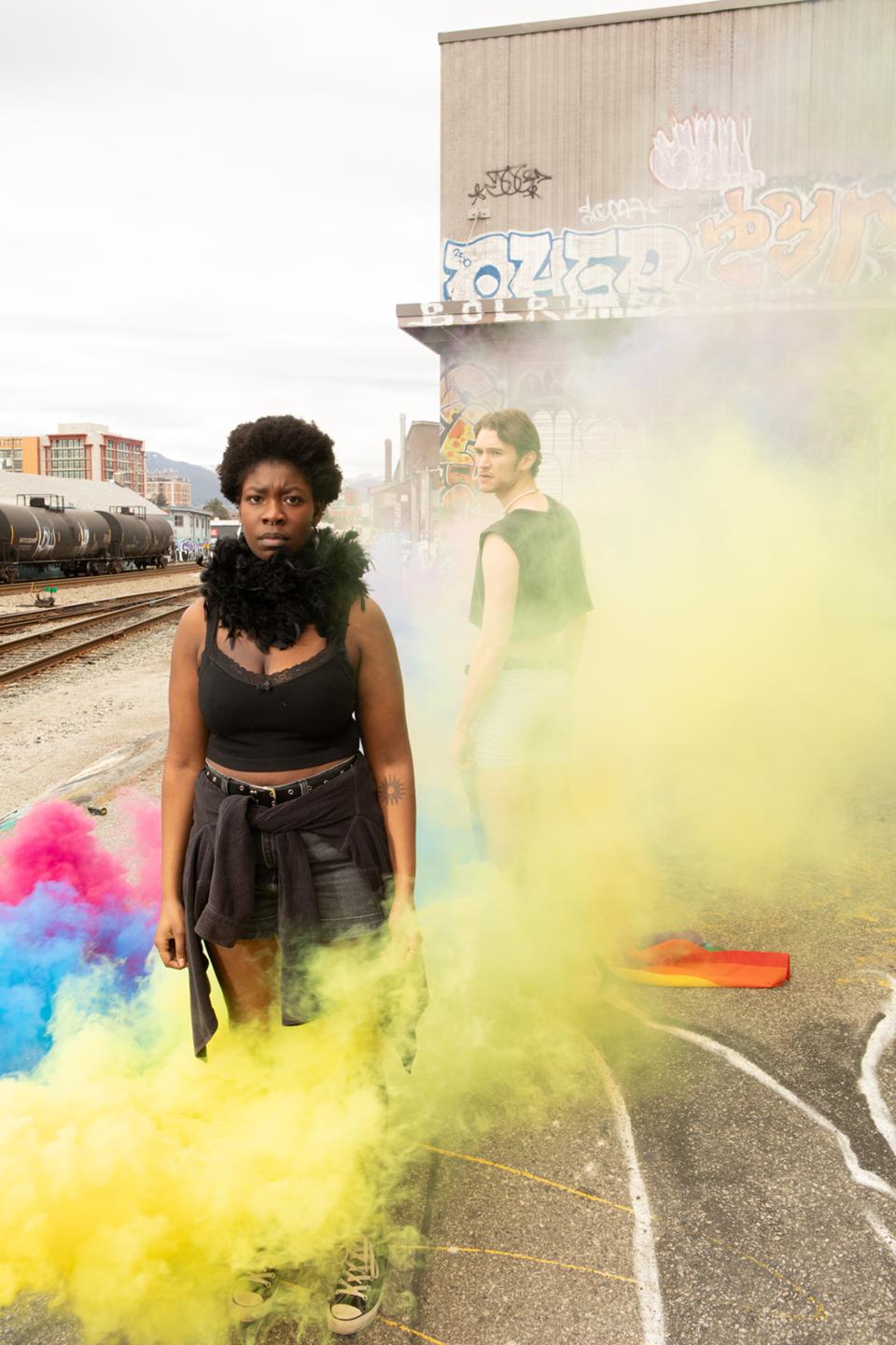 A person sporting an all-black outfit and a black feather scarf stands in an urban landscape including graffiti and train tracks while another person stands behind them, facing the other direction but looking back at them over their shoulder. Yellow smoke and some blue and pink smoke permeate across them. 