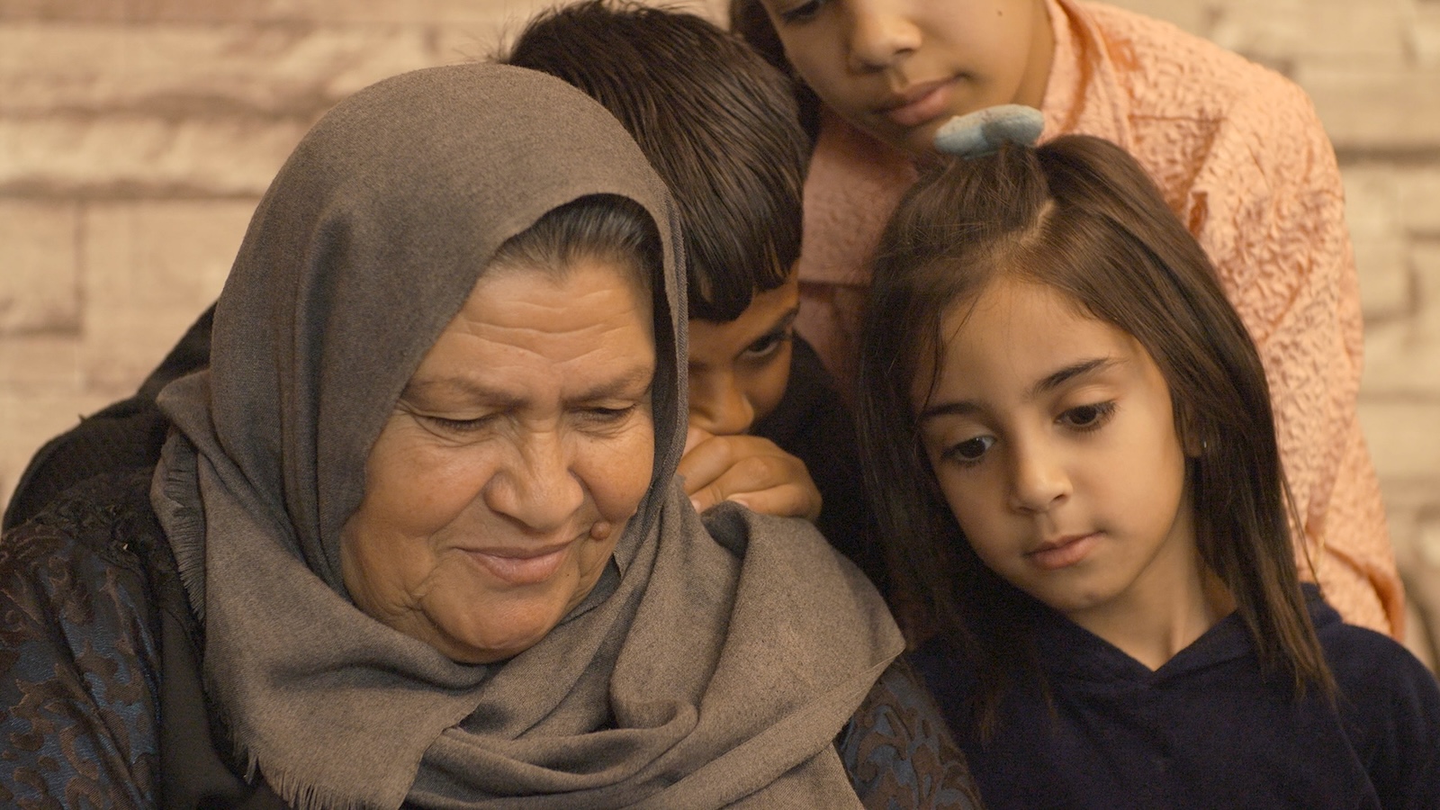 An elderly woman in a taupe headscarf looks down, smiling. She is surrounded by three young children.