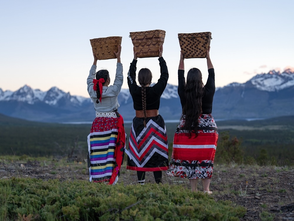 Three women in colourfully patterned skirts stand in a meadow with their back to the camera, holding up Indigenous woven baskets over their heads. Snow-capped mountains can be seen in the distance.