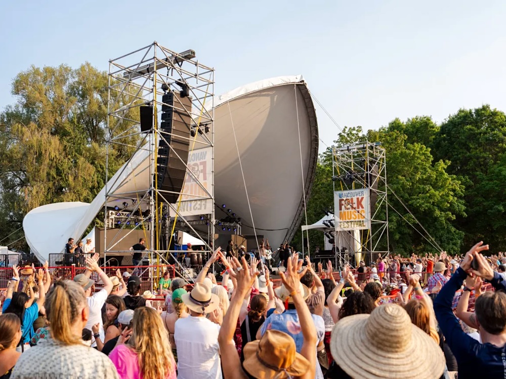 A large crowd of people with arms raised, looking toward an outdoor stage under a white canopy.