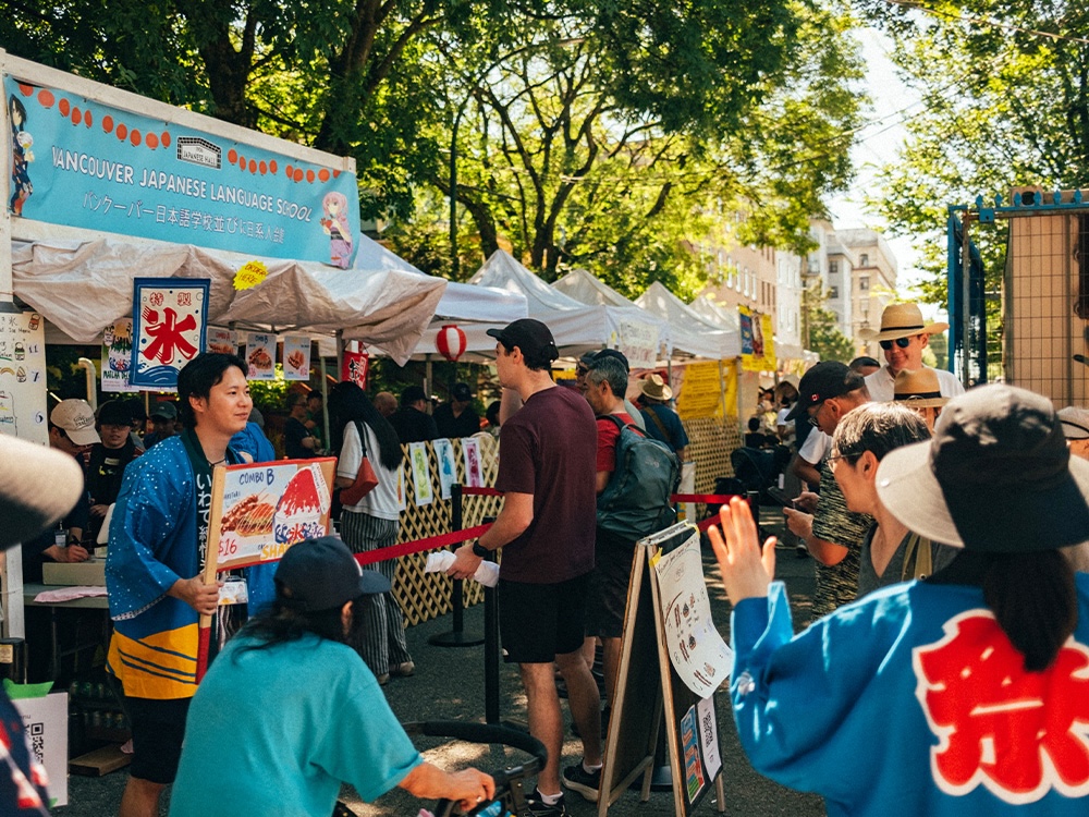 Festival goers line up for kakigori (shaved ice) and other popular treats at the Vancouver Japanese Language School food booth. They are on a shaded street featuring signage for different vendors.