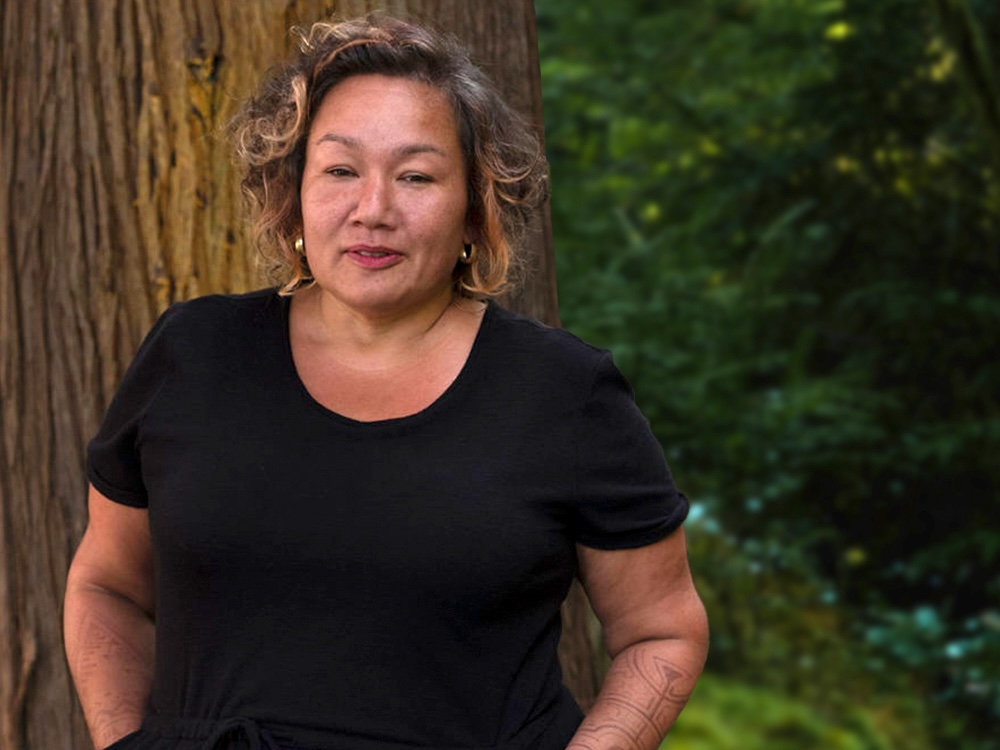A woman with medium skin tone and short wavy brown hair with highlights, wearing a black T-shirt, stands in front of a tree trunk with the forest in the background.