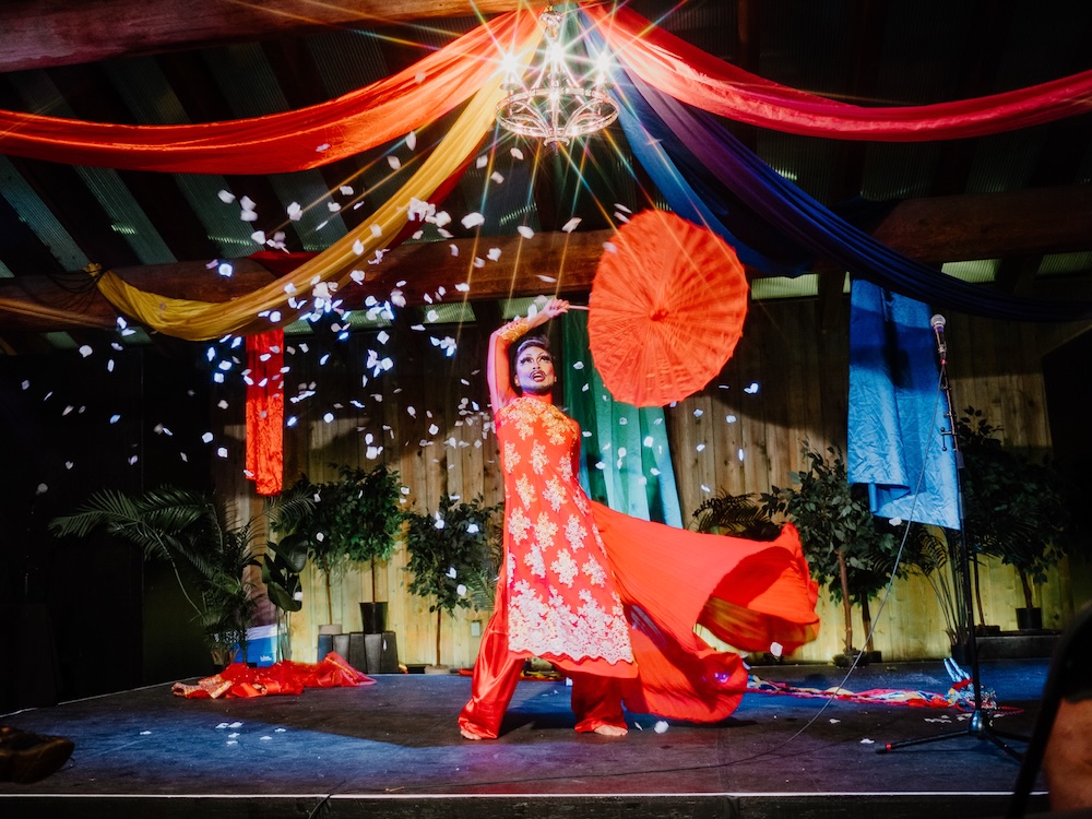 A Fringe performer dressed in all red holds a red umbrella over their head. They are onstage and above them is a chandelier.