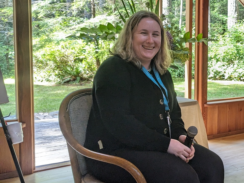 A woman with light skin tone and shoulder-length blond hair sits in a chair with a microphone in her hands on her lap. Behind her is a lush garden of greenery.