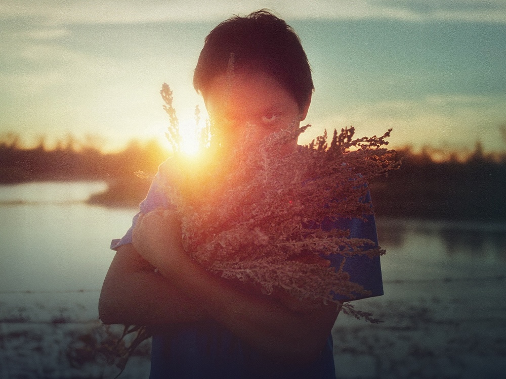 A young boy holds a bunch of plants in his arms and looks up at the camera. Behind him, the sun is setting.