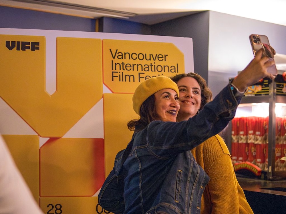 Two filmgoers smile for a selfie in front of a VIFF poster. The person on the left wears a jean jacket and a yellow beret, and the person on the right wears a yellow sweater. To their right is a cart of popcorn bags.