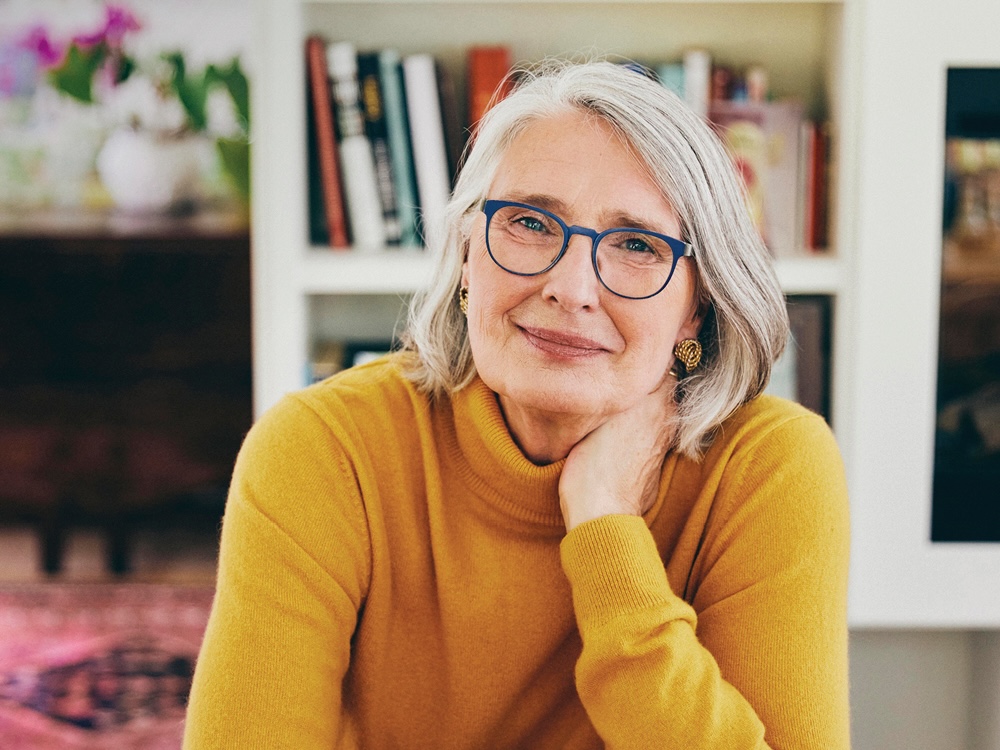 Author Louise Penny smiles warmly at the camera. She wears a yellow turtleneck and blue-framed glasses.