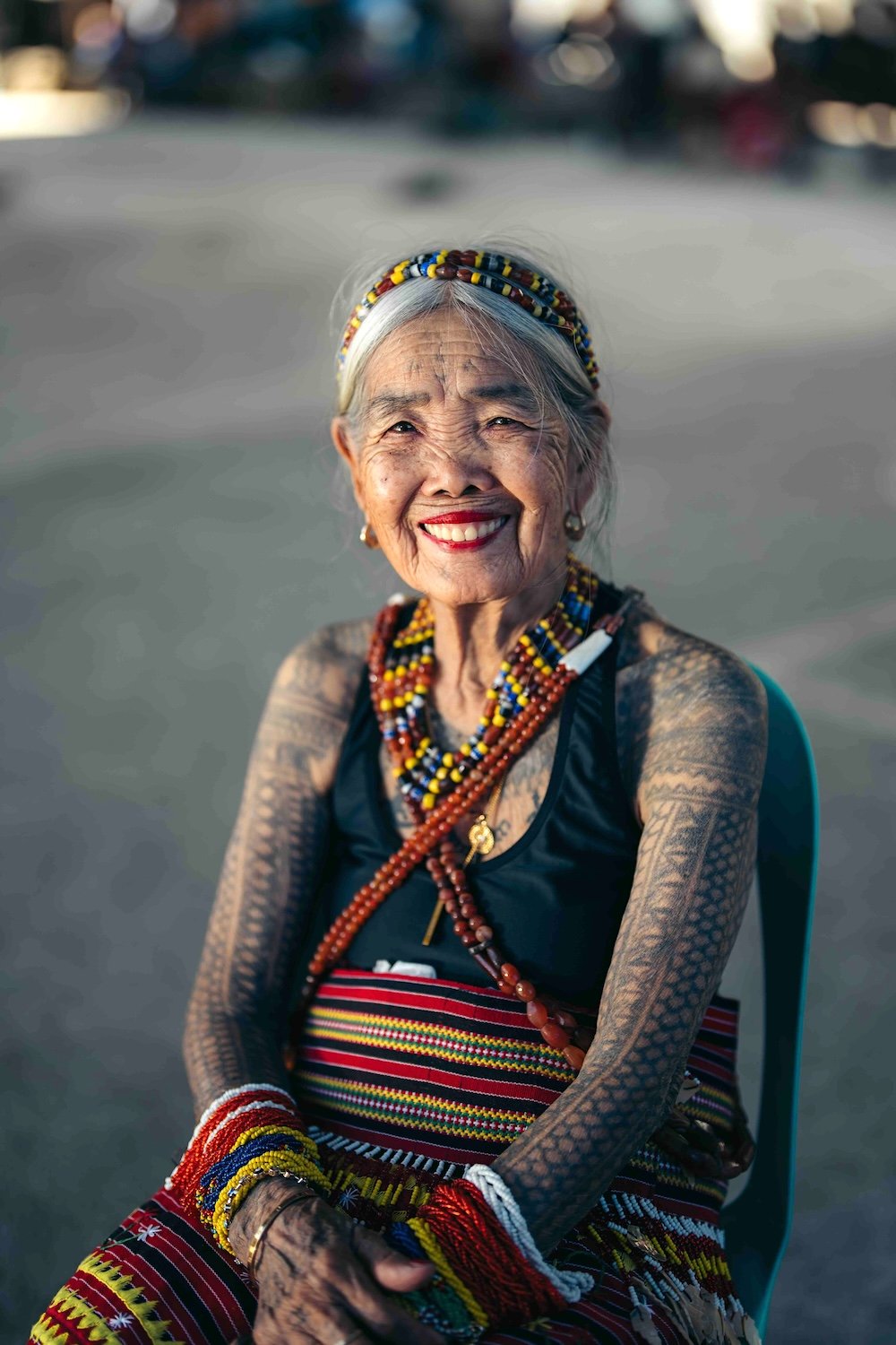 Apo Whang-od is an elderly woman seated on a black chair in an outdoor space, smiling. She has tattoos on her arms and chest, and she wears beads in her hair and across her chest. She is also wearing a colourful wrap around her waist in red, white, yellow and black.