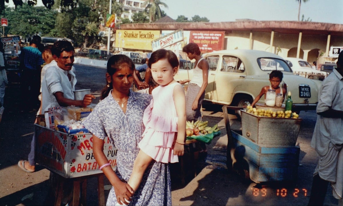 A woman with long dark hair in a braid wearing a blue and white patterned dress holds a young girl with short black hair and a pink dress in the middle of a street lined with market stands selling fruit. People are standing at their stalls or interacting in the background. It’s a sunny day.