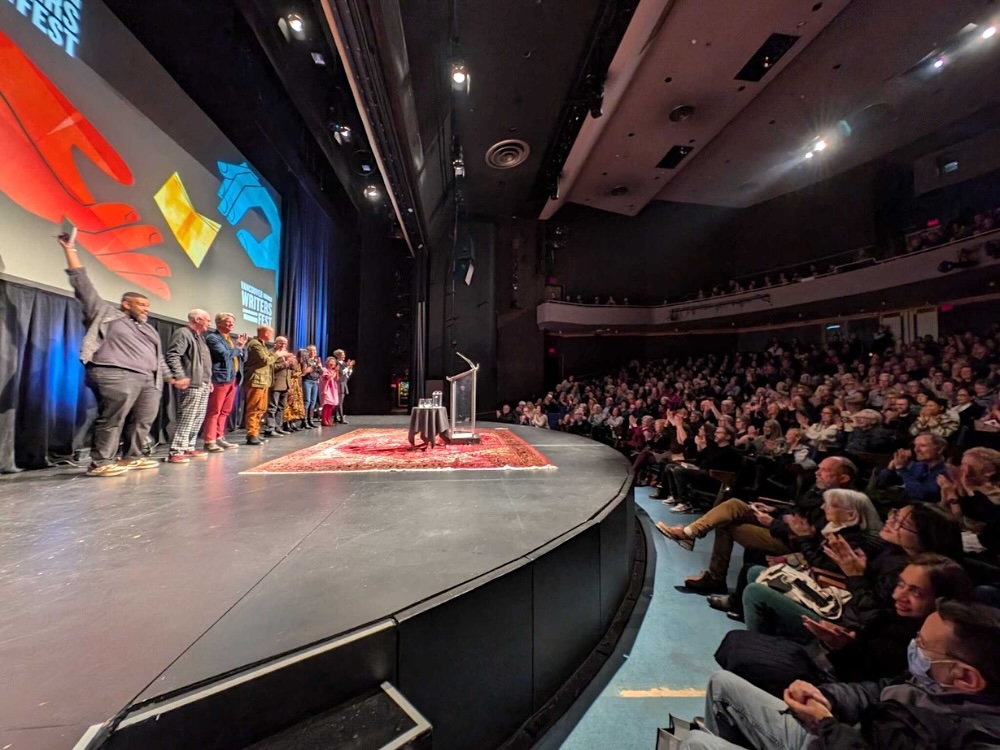 The photo is taken from the side of a stage at a Vancouver Writers Fest event. On the right, audience members clap. To the left, speakers join hands and lift their arms, readying themselves to take a bow.