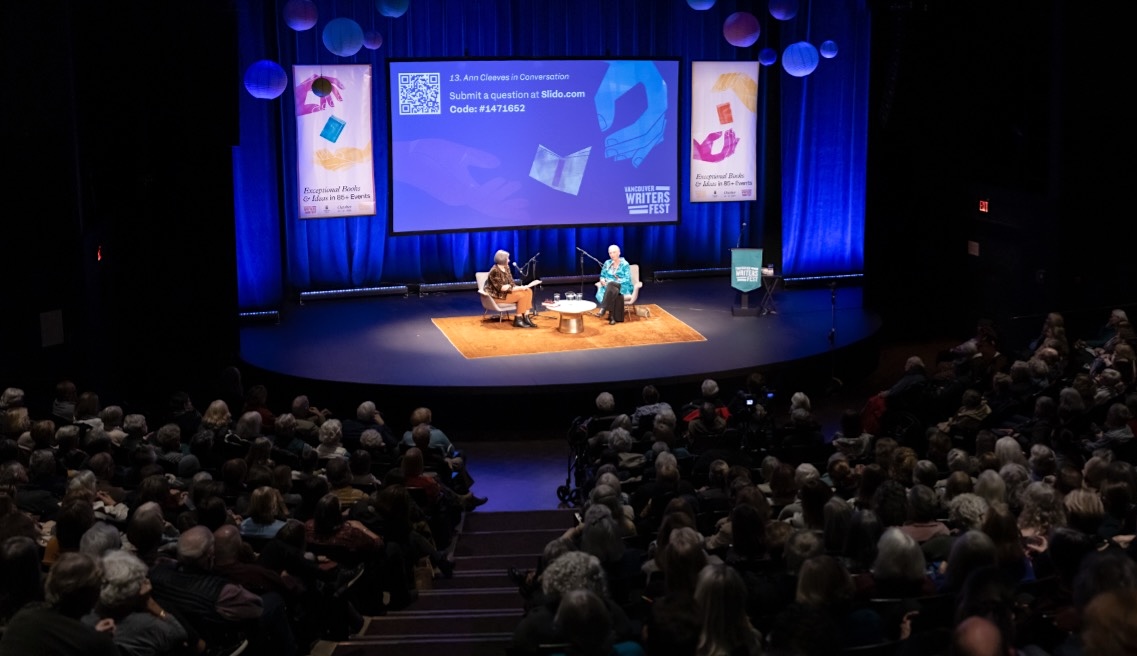 Two women are seated and speaking into a microphone onstage in a full, darkened theatre.