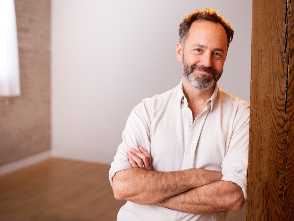 Meditation teacher and author Jeff Warren smiles at the camera. He wears a white button-up shirt and leans against a pillar with his arms crossed. 