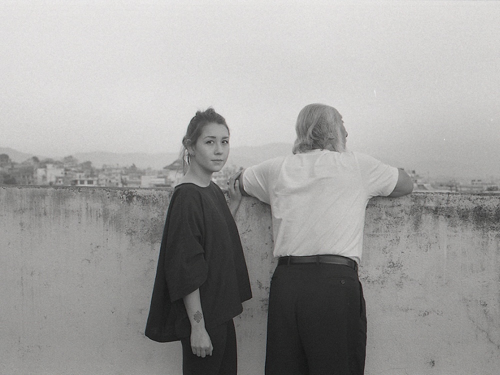 A black and white photo shows two people standing at a short concrete wall overlooking a city. The person on the left is a woman with medium-light skin tone and dark hair tied back, standing sideways and looking at the camera, with her hand on the arm of the person on the right, who has grey hair and their back to the camera, looking out at the view.