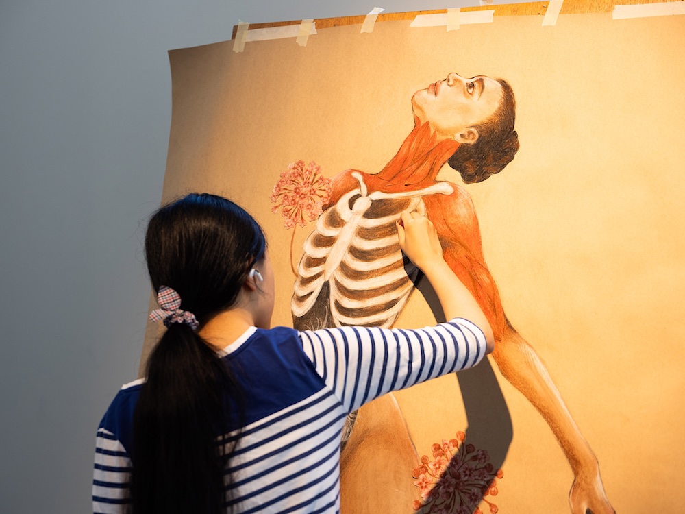 A student in a ponytail is drawing an anatomical illustration of a female body, revealing the underlying muscle groups and rib cage with flowers sprouting out.