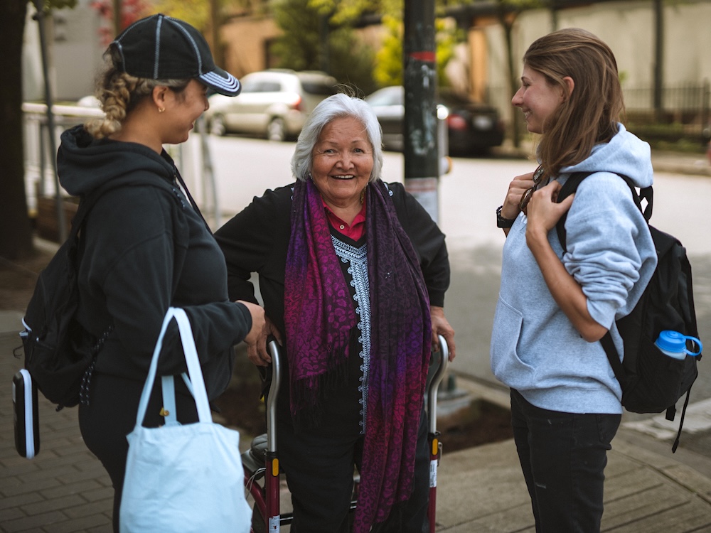 Two young women stand on either side of and face an elderly woman, who is standing with the support of a walker behind her.
