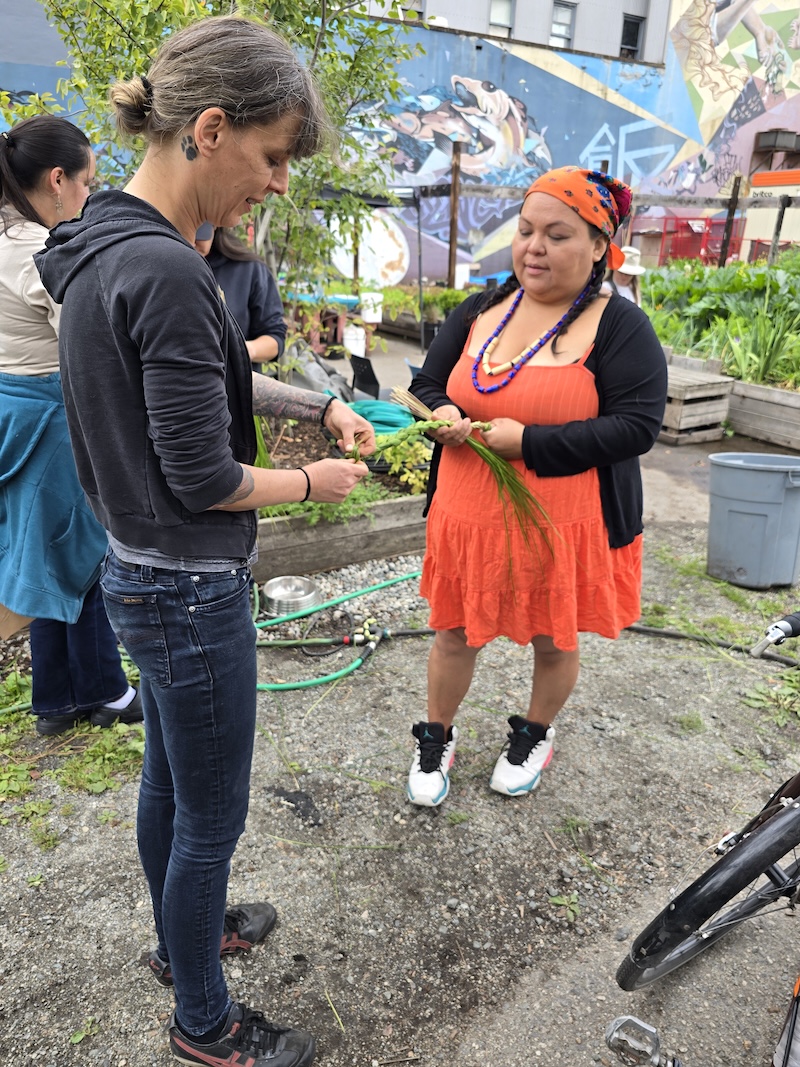 Two people braid grass in the middle of a community garden.