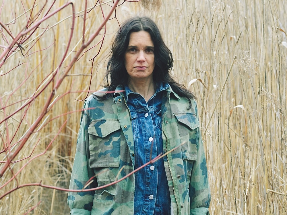 A woman with light skin tone and wavy dark brown hair wearing a camo-patterned jacket stands in a field of tall yellow grass.