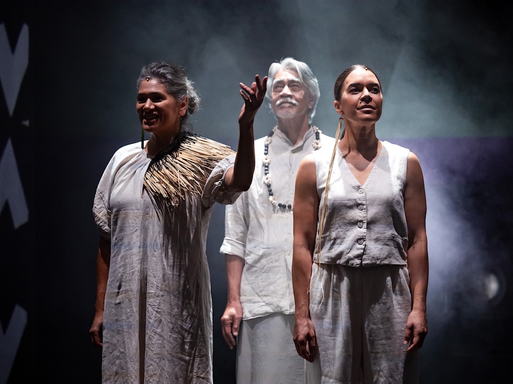 Three performers dressed in all white stand onstage in a triangle formation. They smile at the audience.