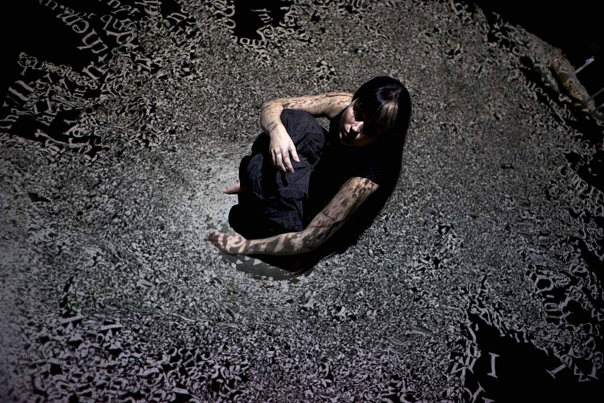A performer sits on a dark stage floor, looking upward at the camera. They sit in the middle of a grey cluster of words that have been projected onto the stage.