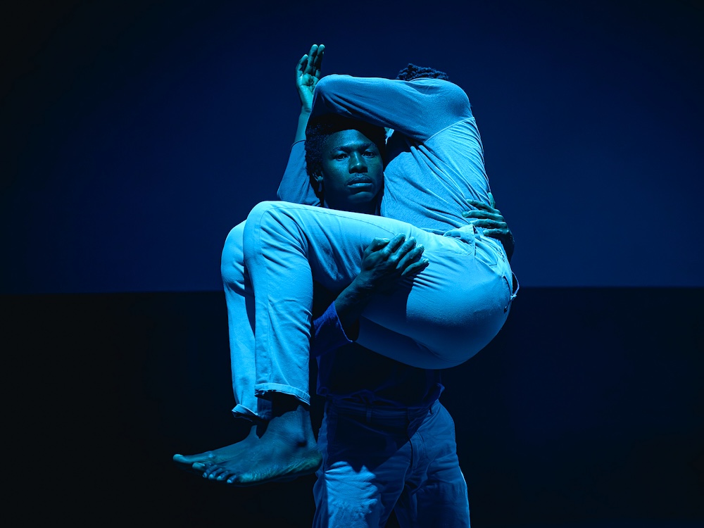 Two performers appear onstage, cast in blue light. One of them looks at the camera, holding the other in his arms.