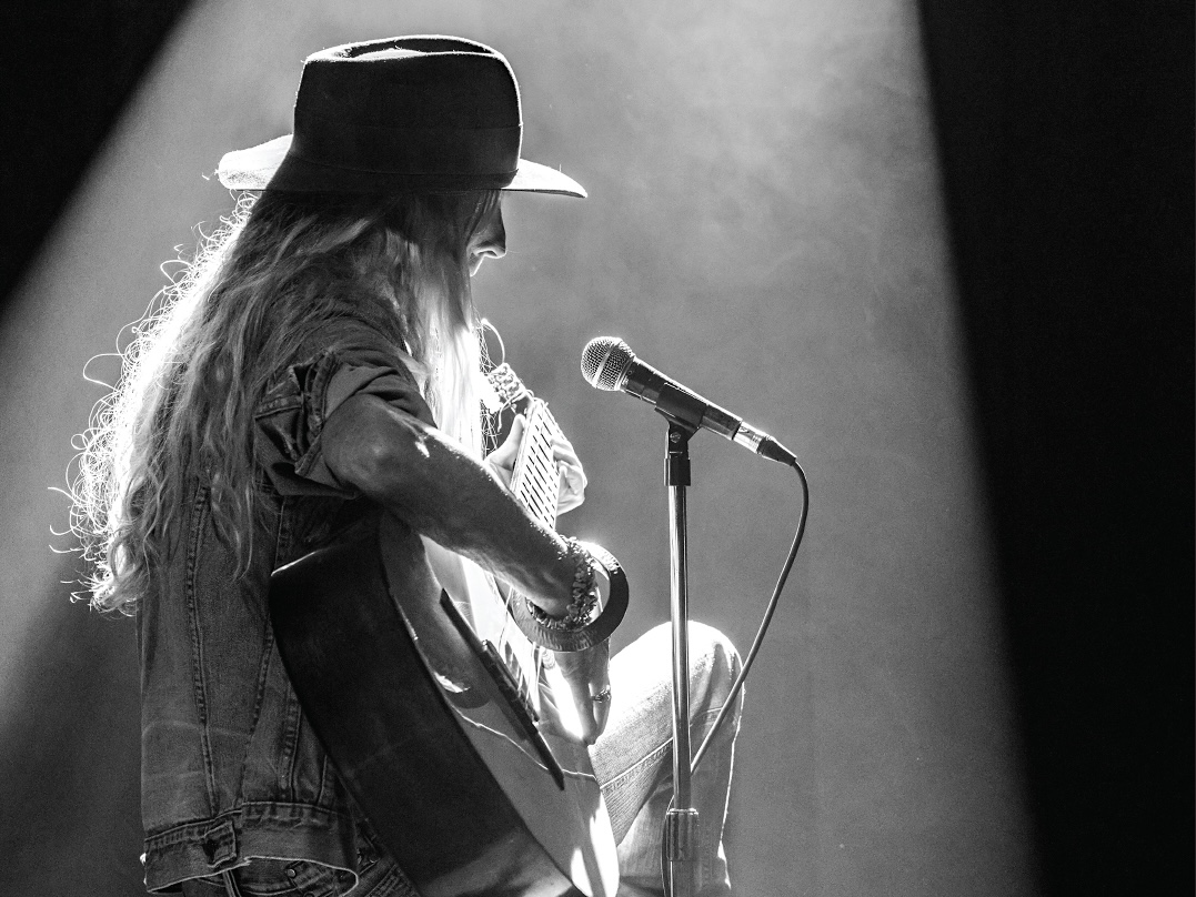 A black and white photo of a performer playing guitar onstage. They sit under a spotlight wearing a hat and jean jacket.