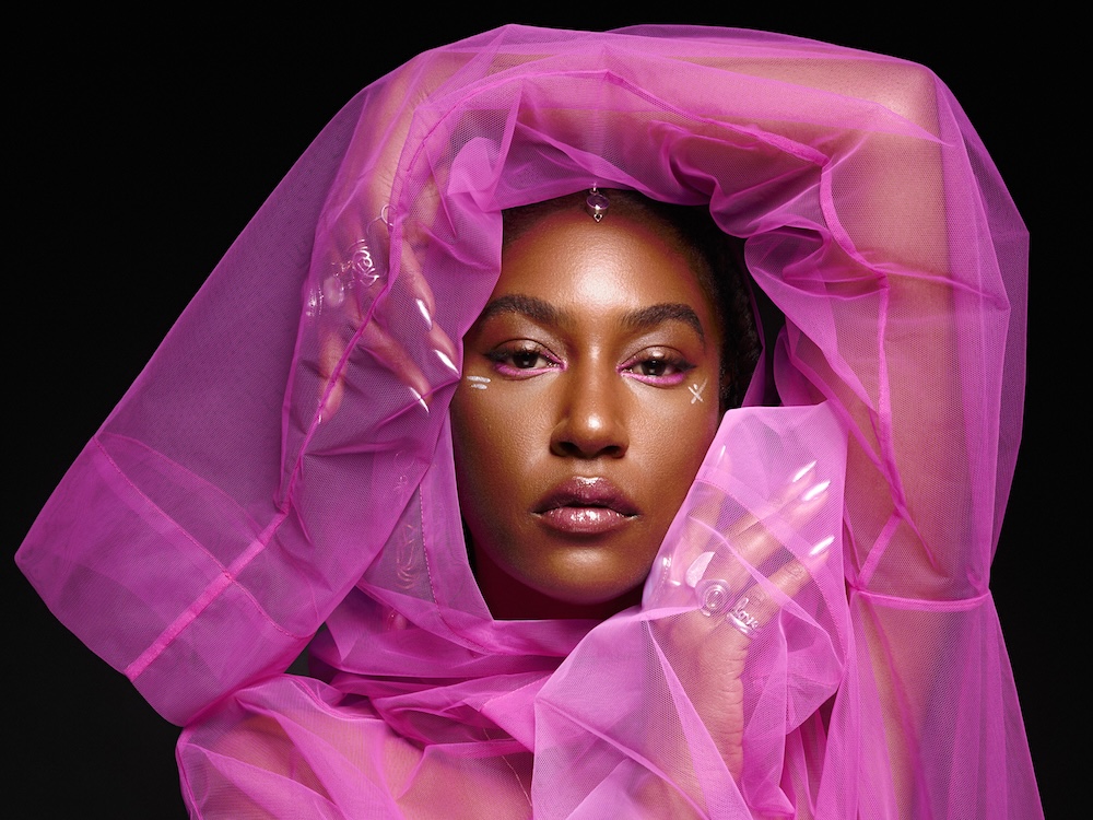 The artist, a woman with medium-dark skin wearing a see-through pink chiffon garment, looks at the camera, framing her face with her hands.