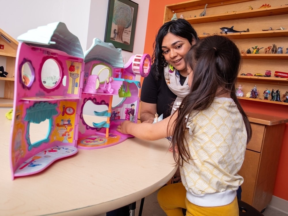 A young girl plays at a bright pink dollhouse with an attentive and kind-looking counsellor by her side. She is in a play therapy room where the walls in the background are full of other toys.