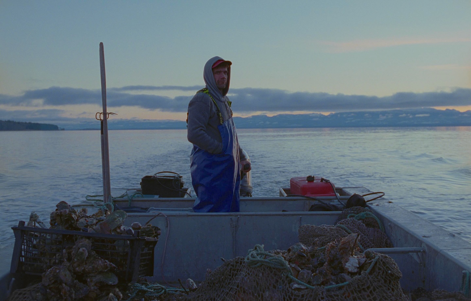 A man stands to the left of the frame in a low white vessel on the water. It&rsquo;s dusk and the sky is dim. He is wearing blue chest waders over a grey hoodie and red ball cap. Around him are nets and other oyster farming gear.