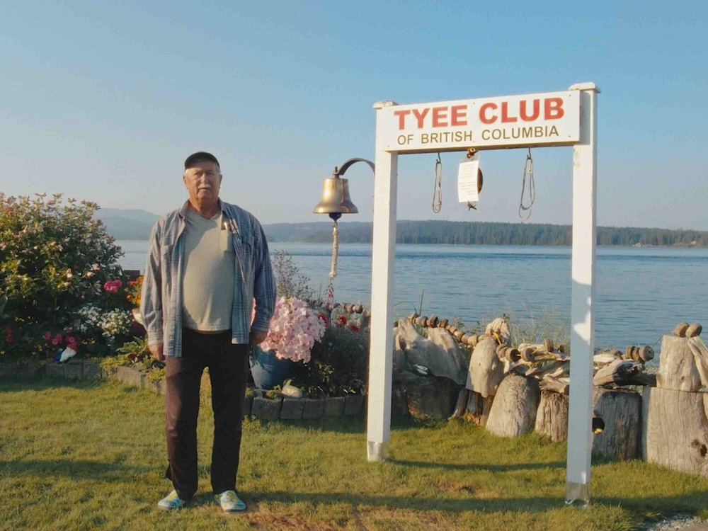 An older man stands in a grassy outdoor area next to the water to the right of the frame. He is wearing a ball cap, a light blue button-down, a light green T-shirt and dark trousers. He stands to the left of a white wooden sign that reads, in red text, &lsquo;Tyee Club of British Columbia.&rsquo; The sign is adorned with a bell, hooks and chains.