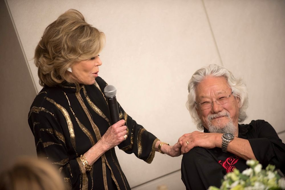 Jane Fonda speaks into a microphone with one hand on a seated David Suzuki's shoulder.