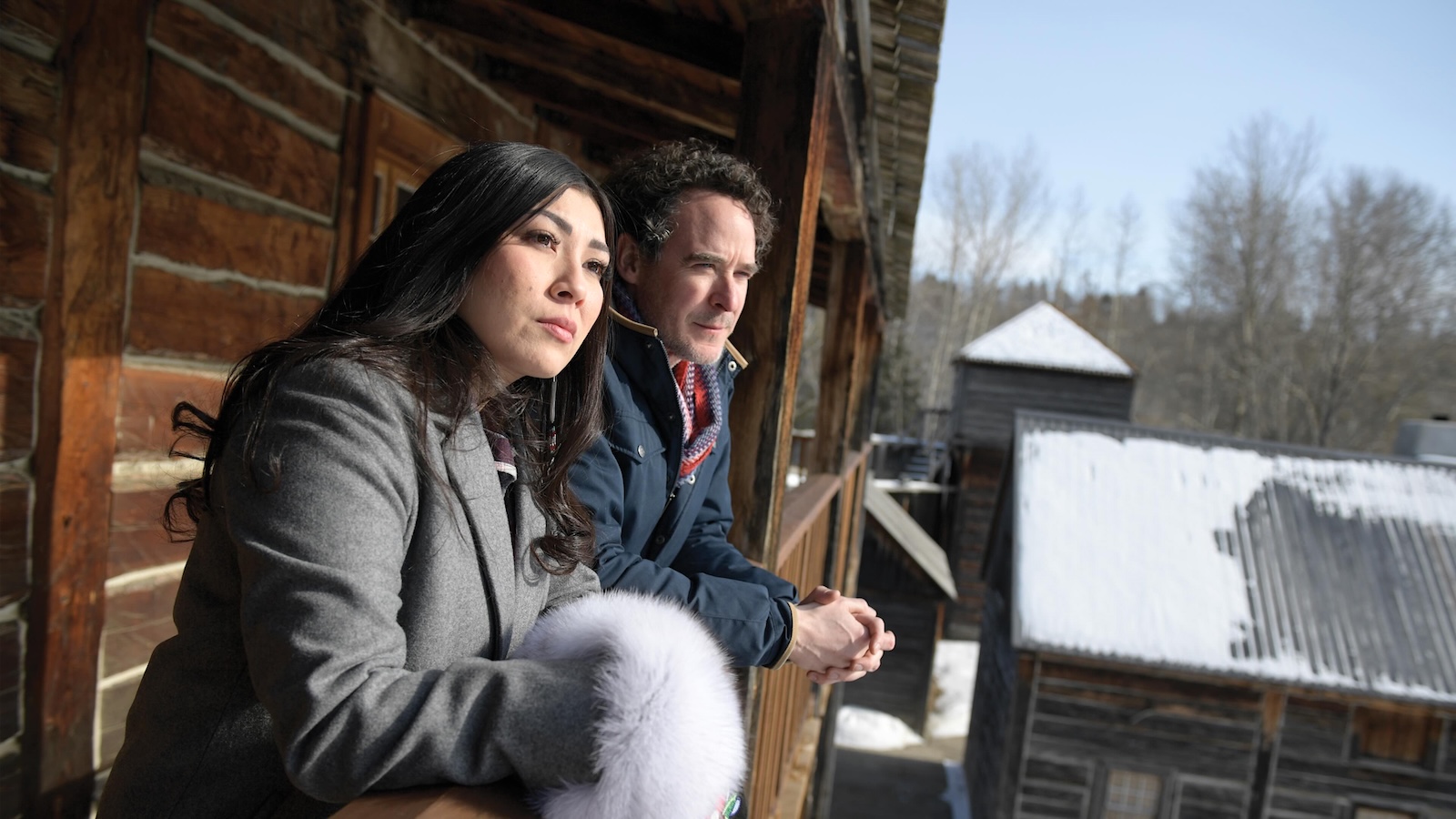 Erin Goodpipe, left, and Saxon de Cocq, right, stand on the balcony of a dark wood building, looking to the middle distance on a clear day. Goodpipe has long dark hair and a grey jacket; she is holding a mauve piece of fur. De Cocq has dark curly hair and a blue jacket over a red shirt.