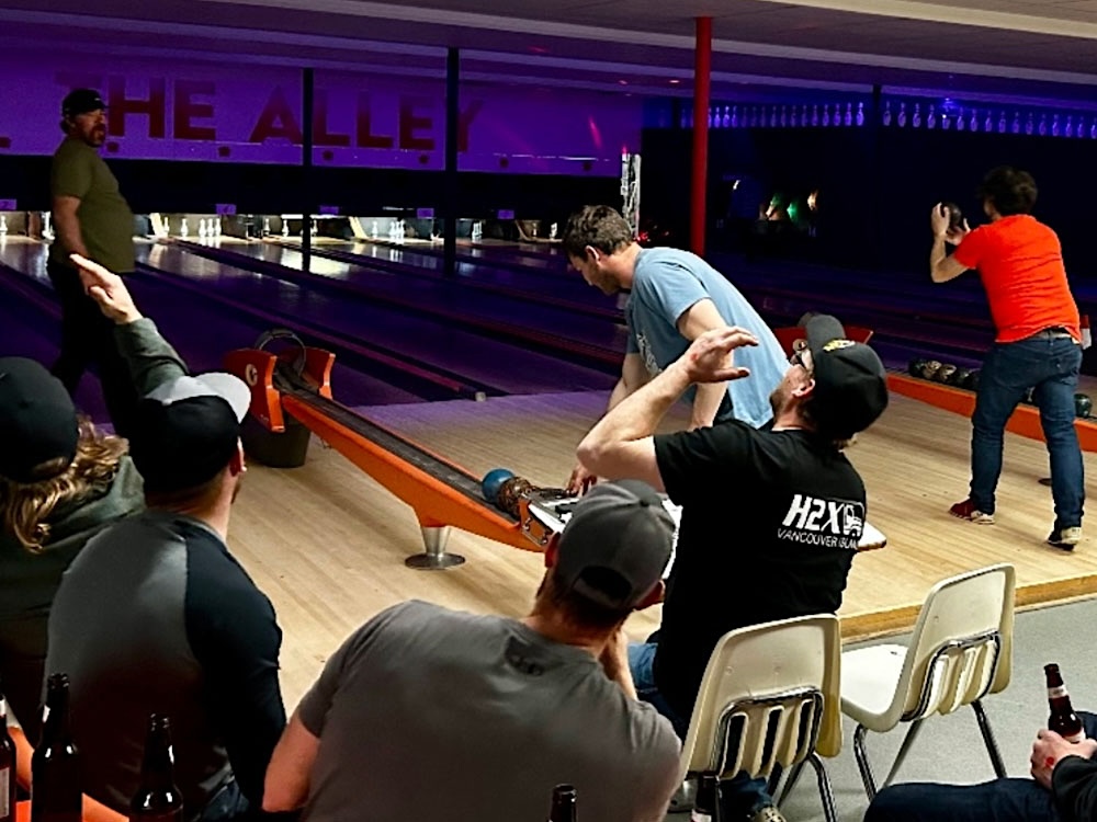 A crowd of people in casual streetwear are seated around a lane at a bowling alley. The lights are dim.