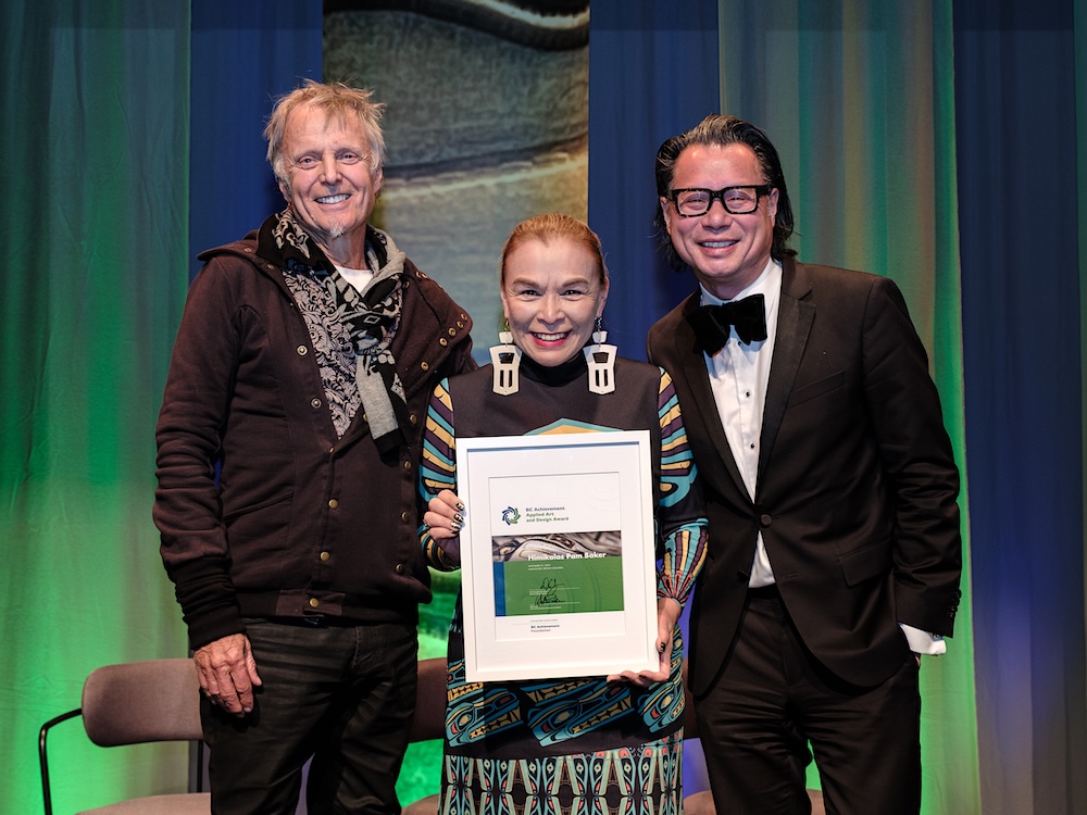 Pam Baker, flanked by John Fluevog and Fred Lee, holds a framed award certificate, and wears a printed dress and bold earrings.