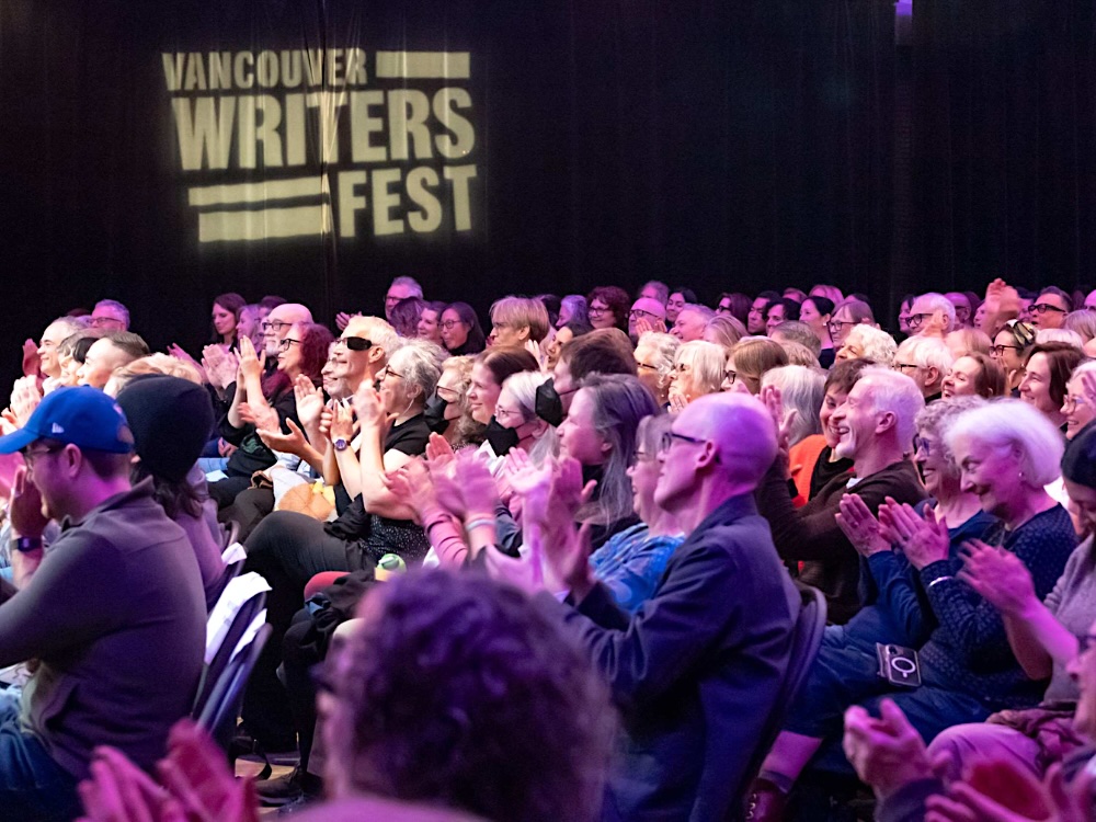 An audience applauds under purple lights with the words &lsquo;Vancouver Writers Fest&rsquo; projected on a wall.