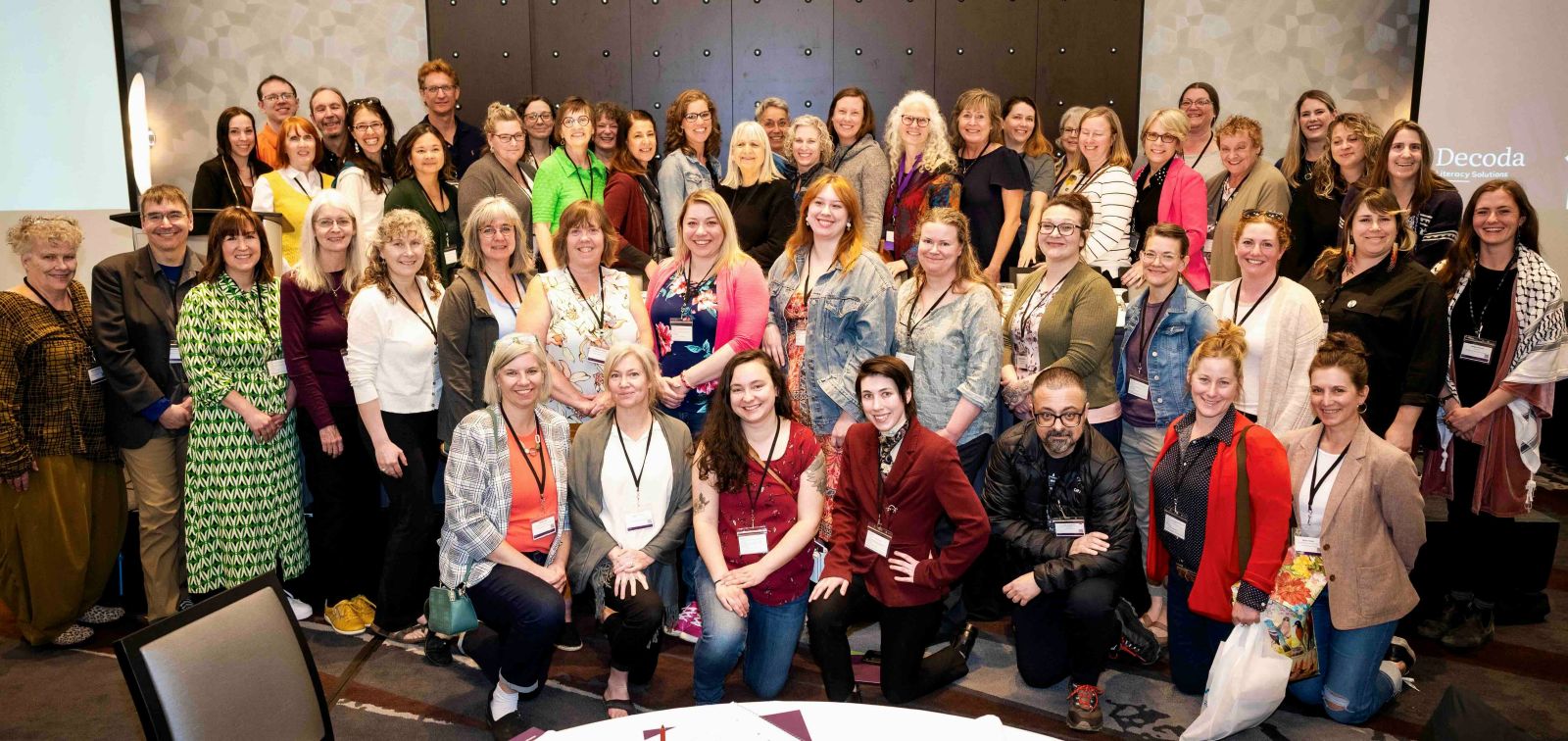 A large group of people poses together in an indoor space for a group photo. They are wearing matching conference lanyards.