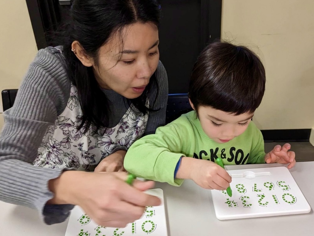 A woman in grey, at left, is seated at a grey table with a young child in green, at right. They are working together to use white plastic sheets with green numbers on them. They are tools that help teach a child how to write numbers.
