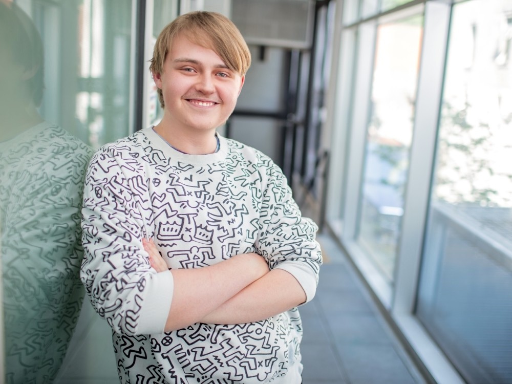 A young man with light skin tone and blond hair stands with his arms crossed and smiles with confidence.