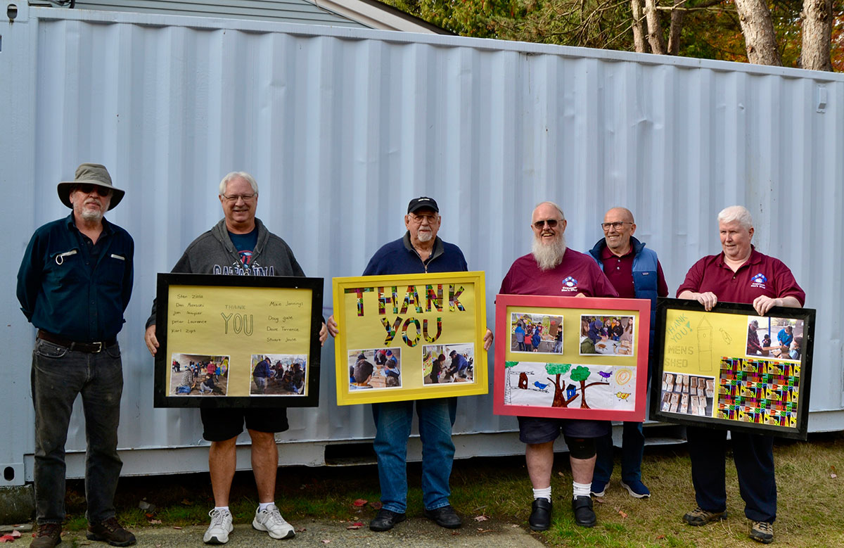 Six members of the Coquitlam Men’s Shed are standing with their backs to a shipping container on a sunny fall afternoon. Four of them are holding large framed collages that have “Thank you” handwritten on them; they are an assembly of thank you cards from students at Edith Cavell Elementary in Vancouver. 