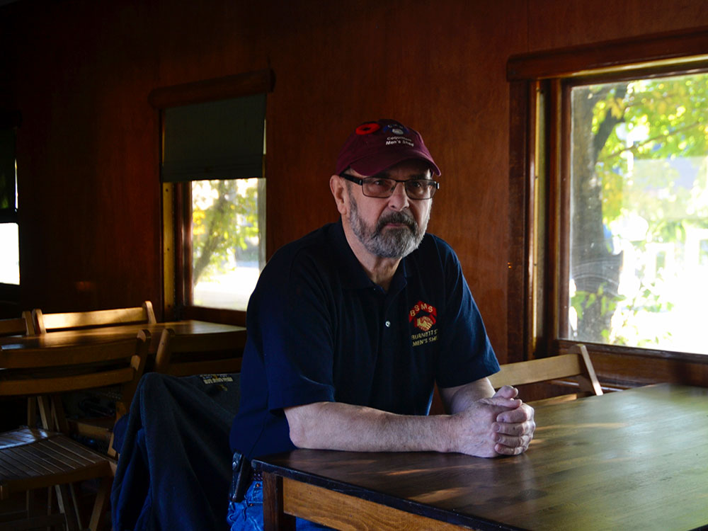 Mike Jennings, president of the Men’s Shed Association of B.C., is wearing a black t-shirt and burgundy ball cap as he sits at a wooden table in a 1921 passenger train car at the Port Moody Station Museum. He has a dark beard and glasses. The Port Moody Men’s Shed is located on the museum’s grounds and the train is used for the collective’s annual general meetings. 