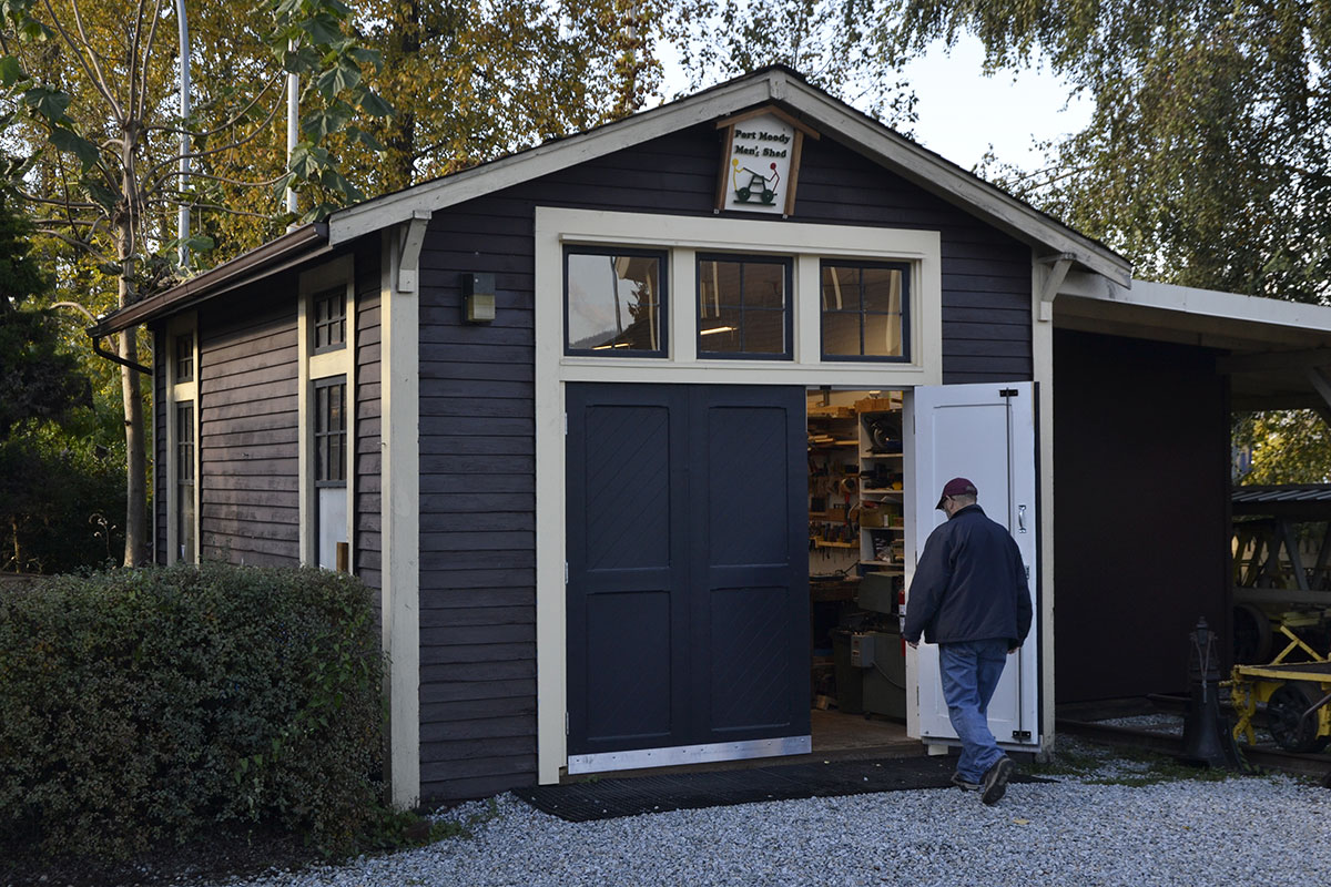 Mike Jennings walks into the Port Moody Men’s Shed on an autumn afternoon. The shed is painted blue with off-white trim. Jennings is wearing a ball cap, a blue jacket and jeans as he walks through an open door painted white. A small handmade sign reads “Port Moody Men’s Shed” above the door. 