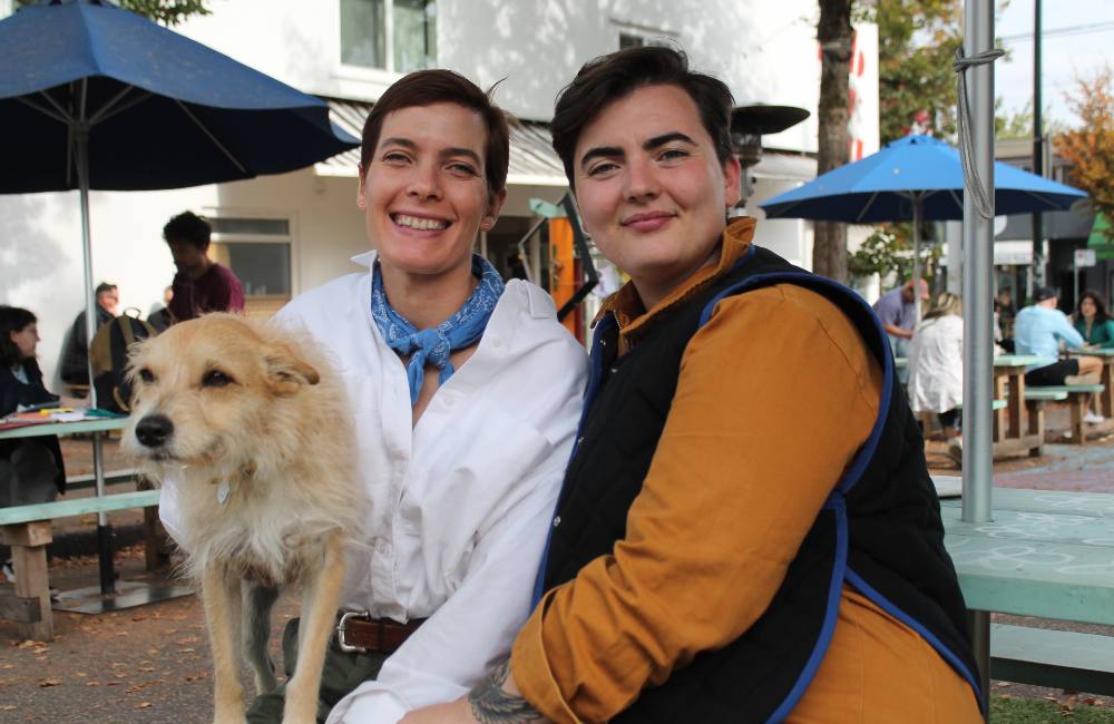 Lizzy Karp, left, and Emma Devin, right, are seated outside at a picnic table with Lizzy’s scruffy terrier, who Karp is holding to the left of the frame. Karp has short dark hair, is wearing a white button-down shirt with a blue scarf and smiling. Devin has short dark hair and is wearing a black vest over a pumpkin-coloured button-down shirt. Behind them is a white building and other people at picnic tables.