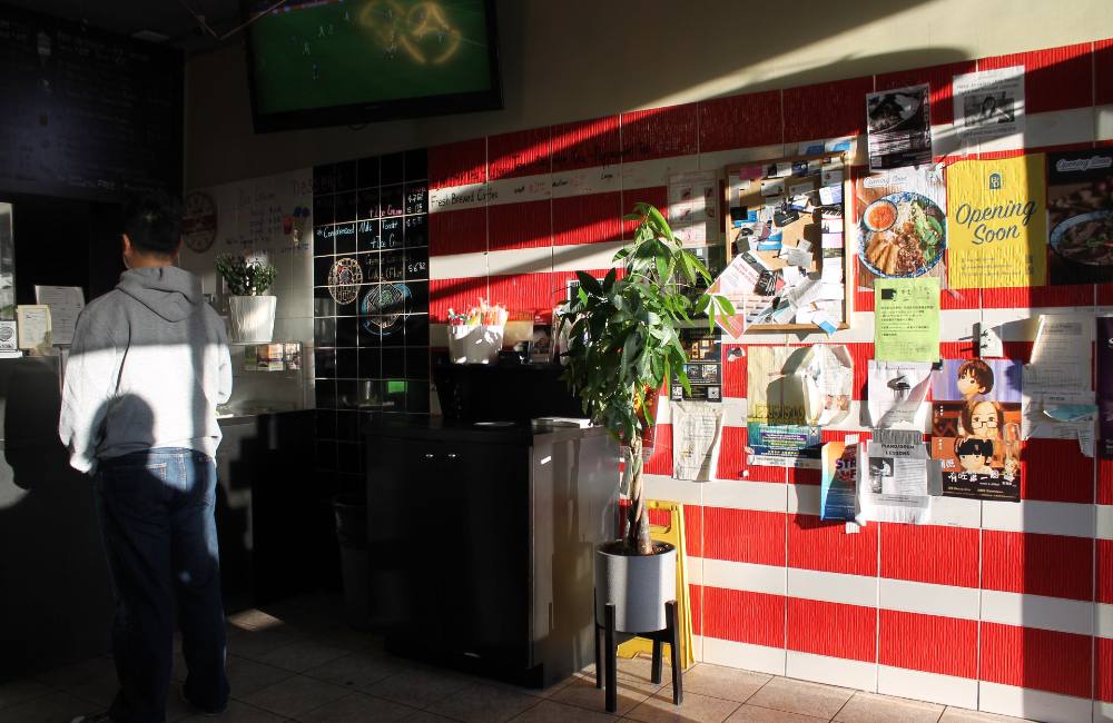 Three stacked photos from the Renfrew-Collingwood neighbourhood in Vancouver depict, from top: a colourful collection of plastic step-stools in front of a storefront; a family of two parents and a toddler walking, shot from the back with restaurant signage that reads “Congee Noodle King” in the foreground to the left of the frame; the interior of an eatery in which one person in grey stands at the counter to the left of the frame while a poster board with a red and yellow sign that reads “Opening soon” is lit with sunlight streaming in from the right.