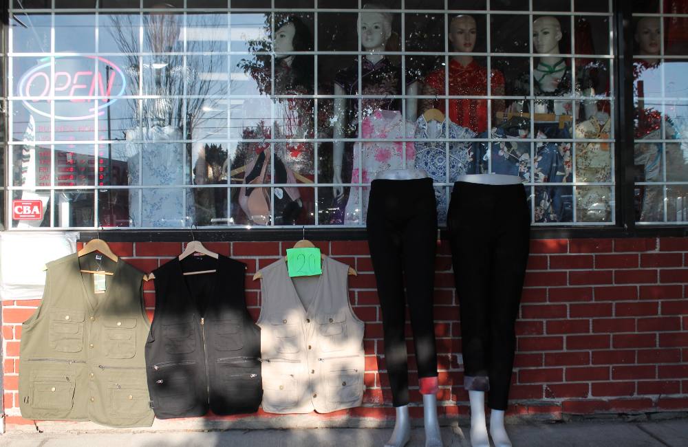 Two stacked photos illustrated two faces of Vancouver. From top, three glassy condo towers near Science World Skytrain station reflect the sunset to the right of the frame. In the bottom image, discounted clothing (three outdoor vests and two pairs of black pants) is arranged on display outside a working-class storefront in the Renfrew-Collingwood neighbourhood.  