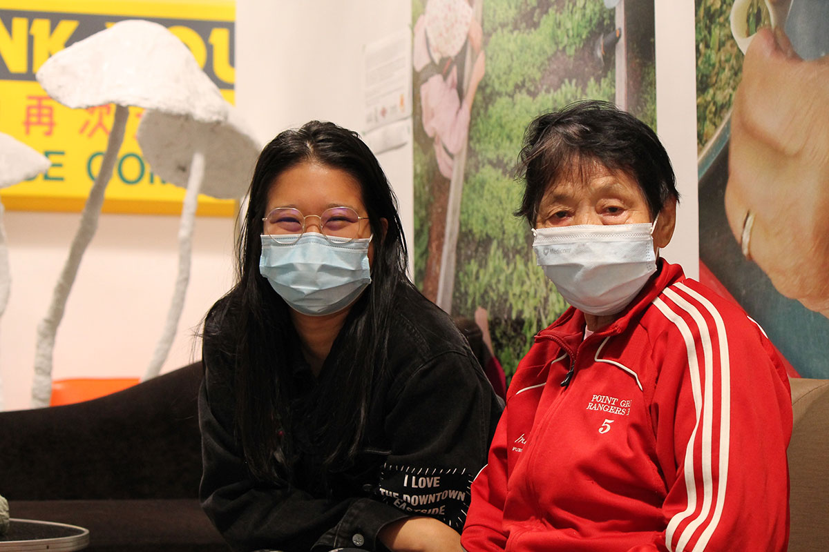 Beverly Ho sits in a medical mask, glasses and black shirt to the left of Yu Rong Li, an elder in a red zip-up track jacket and medical mask. 