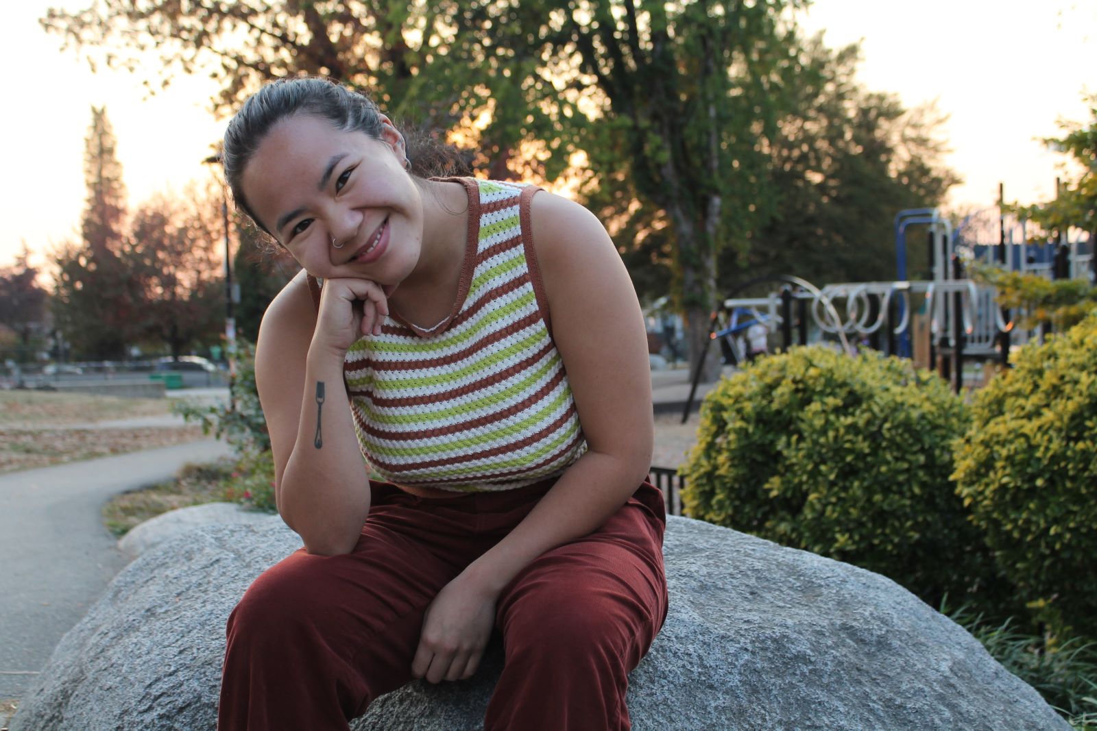 Rachel Lau is seated on a grey boulder in a park. Behind them are bushes, trees and a playground at sunset. They are wearing a green and brown striped sleeveless shirt, brown pants and white sneakers. They are looking at the camera with warmth.