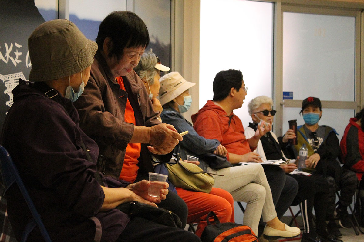 A group of Chinese seniors using walkers and other adults gather in a circle in a large room. Many are wearing medical masks as Nicolas Yung, in the centre of the frame in an orange hoodie and glasses, leads the discussion. 