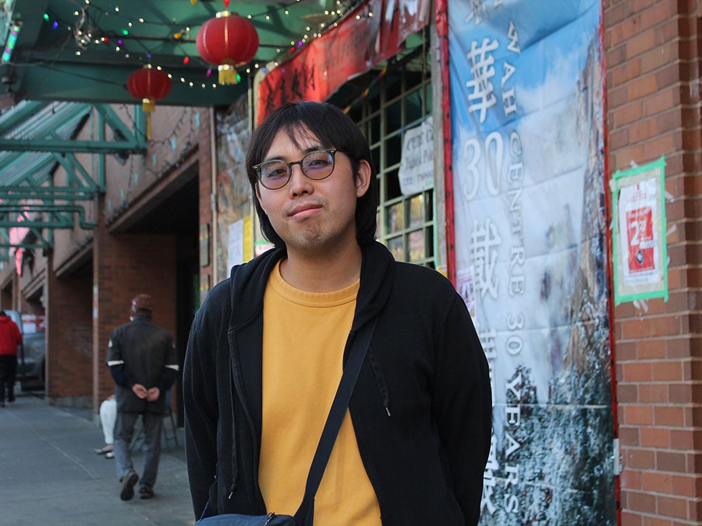Sean Cao, a young Chinese man, stands in front of a building in Vancouver’s Chinatown. A green awning above him is hung with red lanterns and a colourful string of Christmas lights. Cao is wearing tinted glasses and a yellow T-shirt under a black hoodie. He has a soft expression on his face and is standing near a wall with a blue and white mural celebrating 30 years of the Sun Wah Centre.